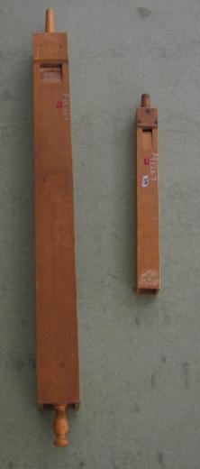 Two square wooden organ pipes of different lengths lying on a gray surface, each with an adjustable stop at the top and a turned wooden finial at the bottom