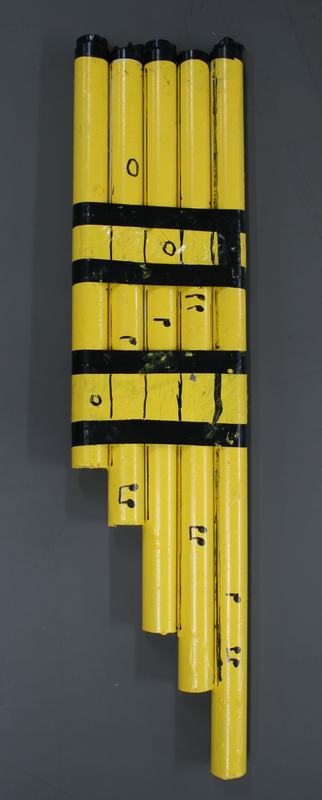 Yellow PVC panpipes with black tape bands and endcaps, five graduated tubes with hand-written musical note labels