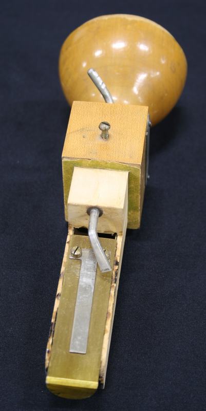 End-on view from the bell side showing the metal reed strip in the wooden channel and the pitch-control lever arm above it, with the turned wooden bell behind