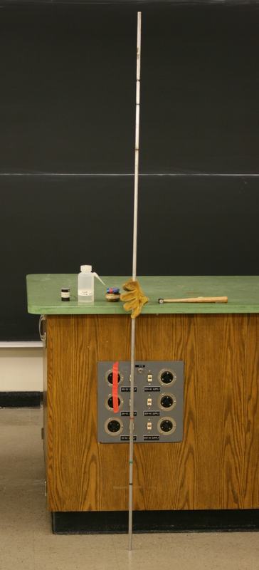 Long metal singing rod leaning against a lecture demo table with a red band visible near the midpoint; leather glove, rosin bottle, and wooden mallet rest on the green countertop
