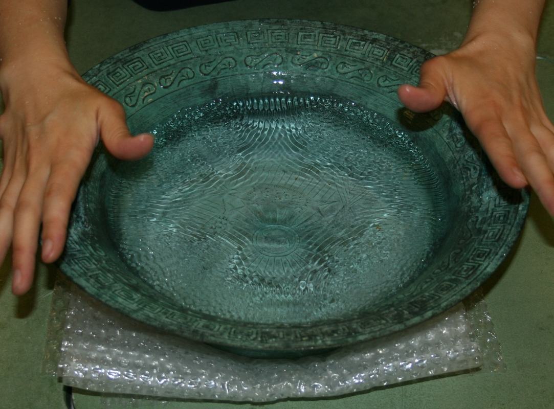 Hands rubbing brass handles of the singing bowl, water surface showing standing wave ripple patterns