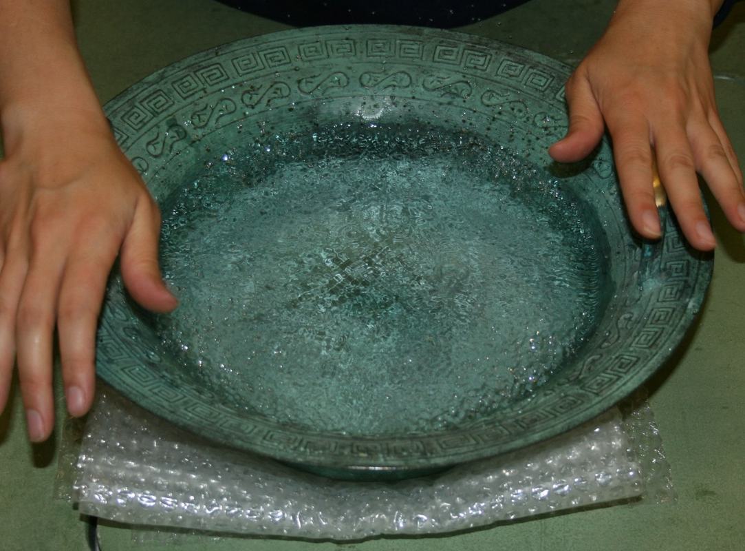 Hands rubbing bowl handles with water showing vigorous splashing and fountain effect from higher-mode excitation