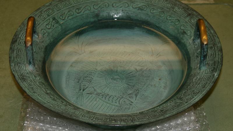 Green patina bronze singing bowl with Greek key pattern on rim, two brass handles, filled with water, sitting on bubble wrap