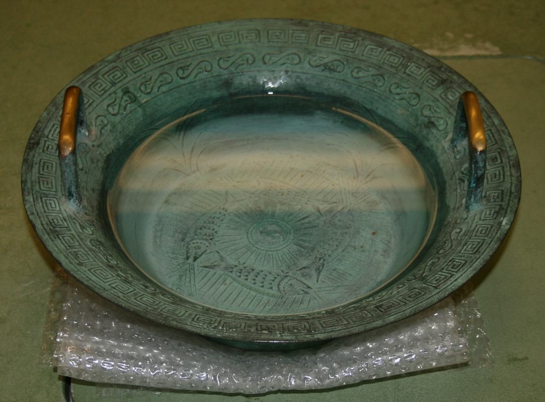 Green patina bronze singing bowl with Greek key pattern on rim, two brass handles, filled with water, sitting on bubble wrap