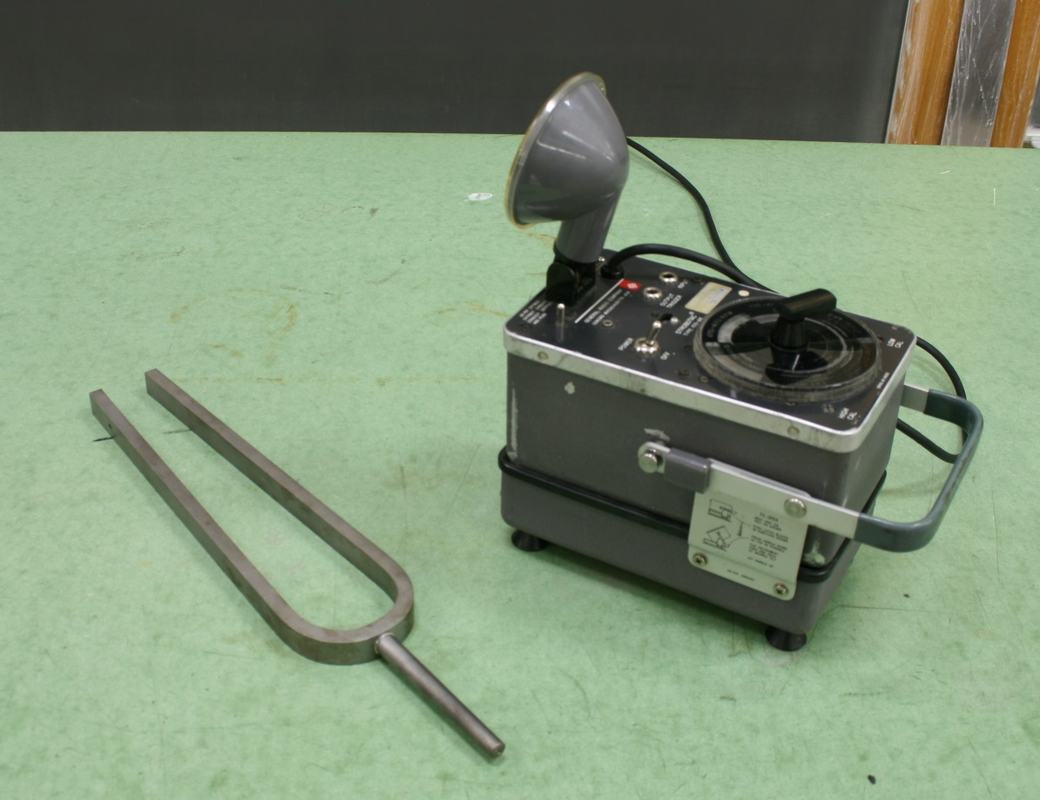 Large metal tuning fork with cylindrical handle lying next to a portable strobe light unit with adjustable dial and reflector