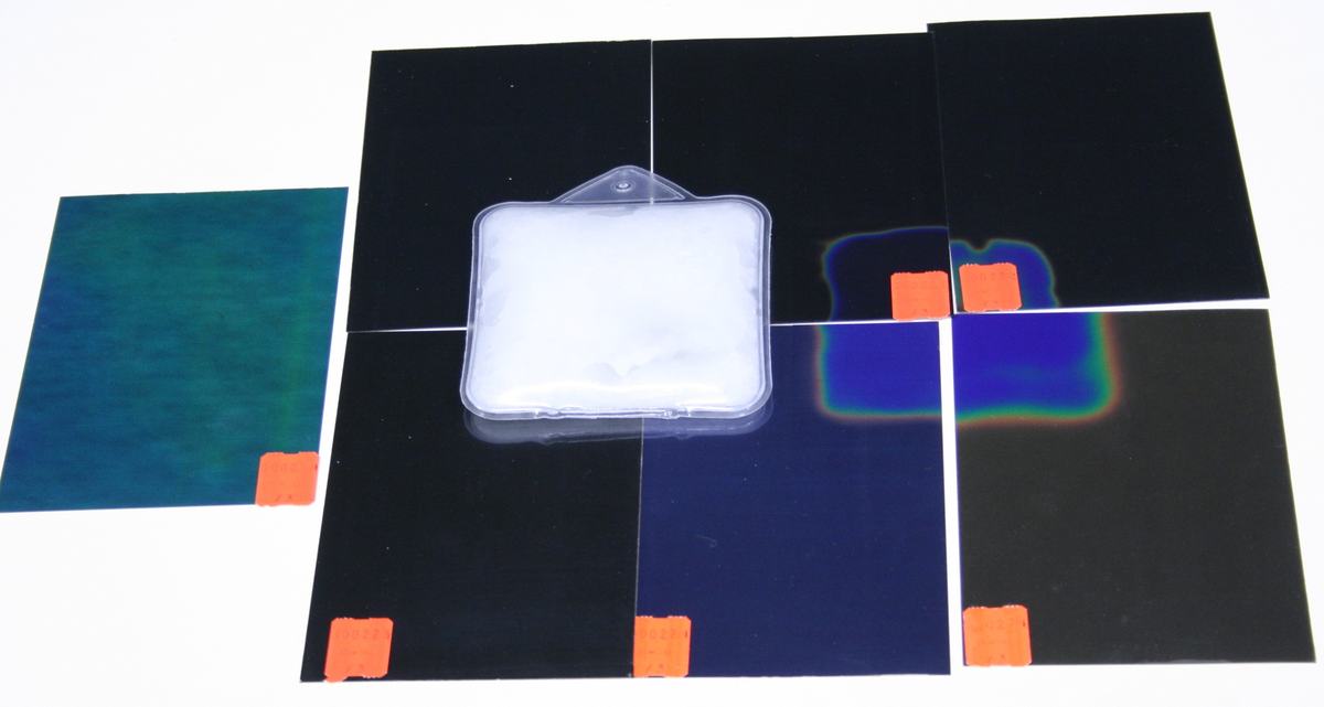 Six liquid crystal sheets on a white surface with a reusable heat pad in center; one sheet shows teal/green activation, two show partial blue activation near the heat pad, others are black; orange identification stickers on corners