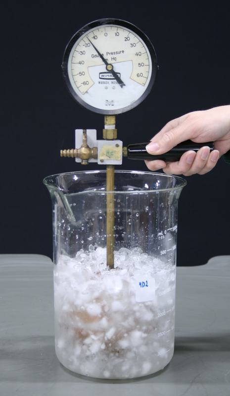 Gas thermometer bulb submerged in ice-water mixture in a large glass beaker, hand holding handle, gauge reading near -20 cm Hg