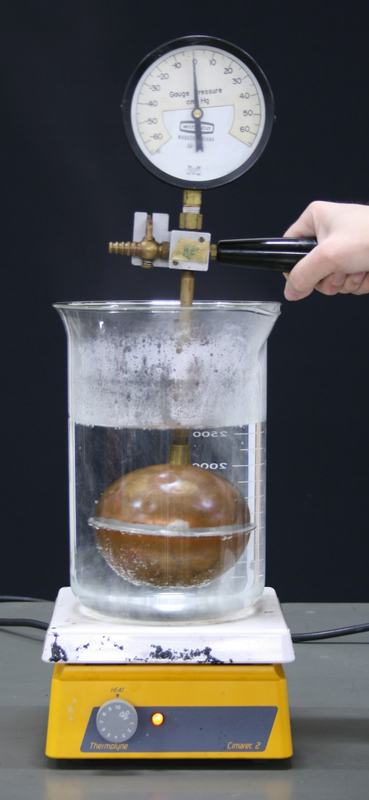 Gas thermometer copper bulb submerged in boiling water in a large glass beaker on a Thermolyne hot plate, hand holding the black handle