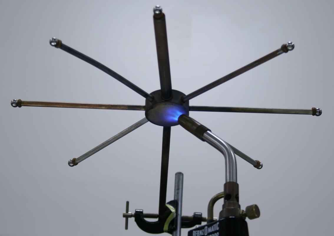 Eight metal rods of different metals radiating from a central hub, with steel balls waxed onto the tips, mounted on a ring stand with a Bernzomatic propane torch heating the hub from below