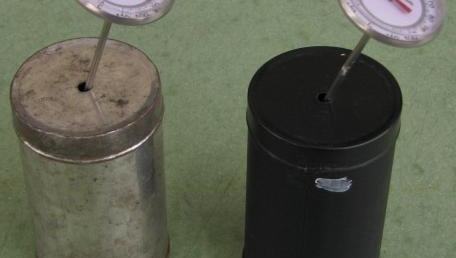 Two cylindrical cans with lids and dial thermometers inserted through the top: a silver/metallic can (left) and a black-painted can (right)