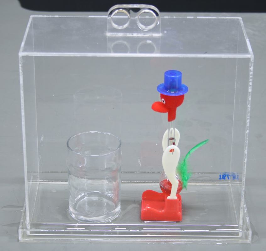 Drinking bird standing upright inside a clear plexiglass case on an acrylic tray, with a glass beaker of water positioned for the beak to reach when tipping