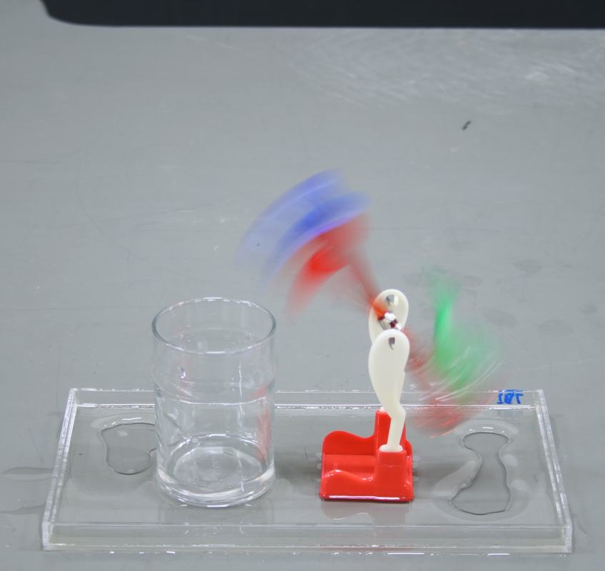 Drinking bird mid-cycle, captured with motion blur showing its rocking oscillation, on a clear acrylic tray with an empty glass beaker beside it