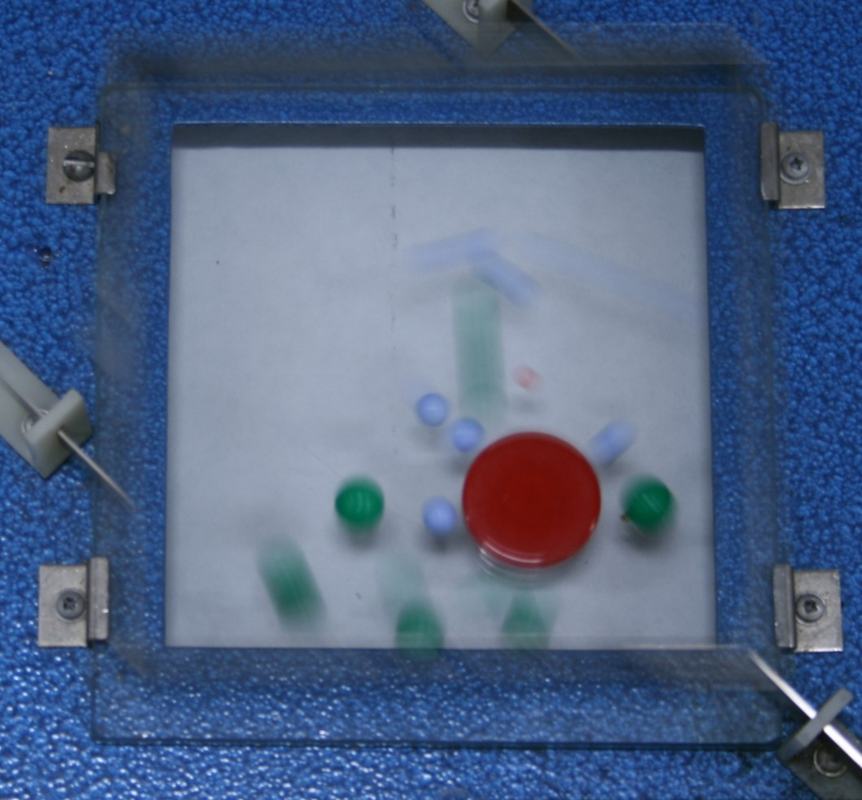 Overhead view of molecular motion demonstrator: transparent square frame with corner clamps on a white glass plate, containing a large red disk surrounded by smaller blue and green spheres, several blurred from motion