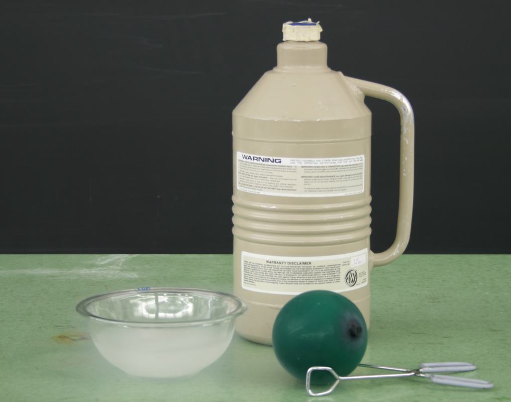 Tan liquid nitrogen dewar with warning label, clear glass bowl, inflated dark green balloon, and metal tongs on a green benchtop