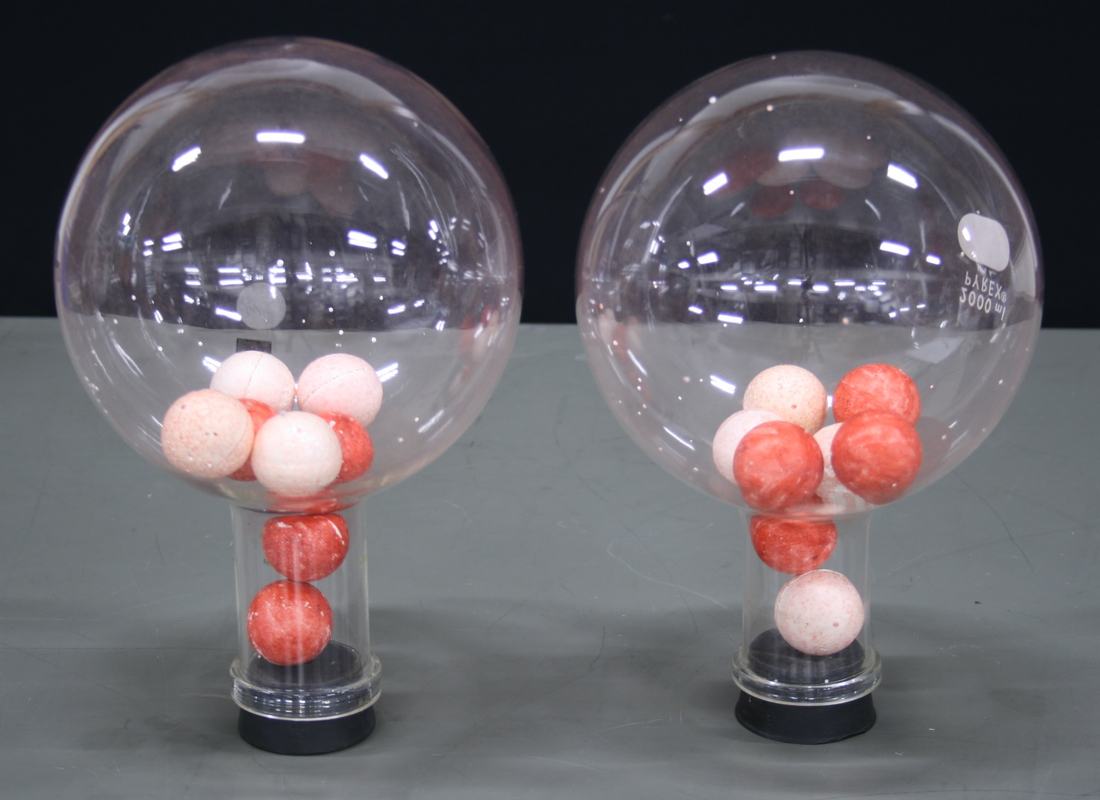 Two large round-bottom glass flasks on clear cylindrical bases, each containing four white and four red foam balls. The balls sit mixed in the flask bodies and necks.