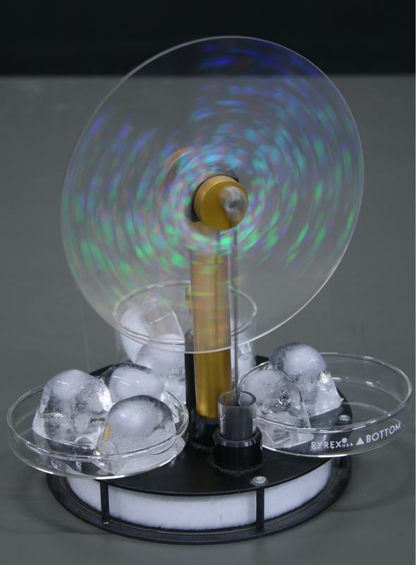 Stirling engine running with three clear dishes of ice cubes placed around the base, flywheel spinning with motion blur