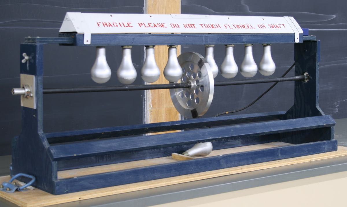 Radiant heat engine: blue wooden frame with eight large frosted incandescent bulbs mounted in a row above a horizontal thin aluminum tube (shaft) passing through a perforated metal flywheel disc. A white sign reads "FRAGILE PLEASE DO NOT TOUCH FLYWHEEL OR SHAFT."