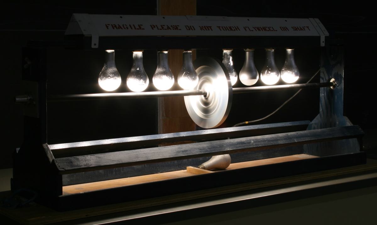 Radiant heat engine in operation: bulbs glowing brightly, illuminating the aluminum shaft. The flywheel disc shows motion blur from rotation. Dark background emphasizes the lit bulbs.