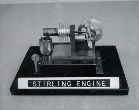 Small Stirling engine on a black base with "STIRLING ENGINE" label. The engine has a finned displacement cylinder on the left, a power cylinder with connecting rod in the center, and a flywheel on the right. An alcohol burner sits beneath the displacement cylinder.