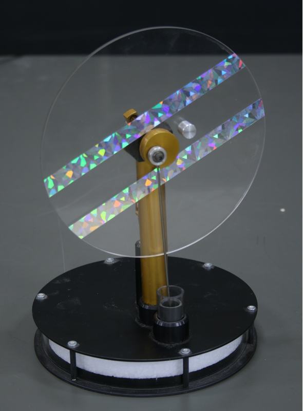 Visible Stirling engine at rest: clear acrylic flywheel disc with two diagonal holographic tape strips, gold brass displacer cylinder, small gray power piston at the base, mounted on a black plate with white foam displacer visible through the side