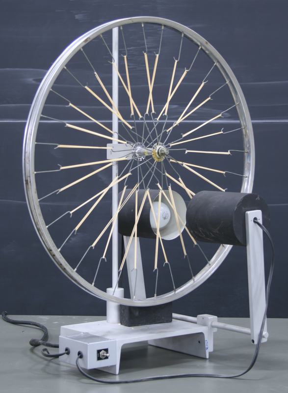 Bicycle wheel mounted vertically on a white stand with rubber bands stretched from an offset center hub to the rim. A large black cylindrical heat lamp is positioned to one side, plugged into a switch box on the base.