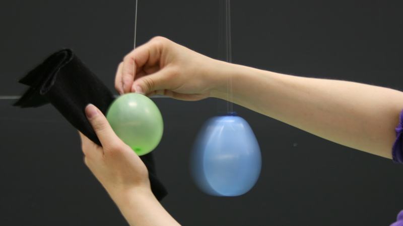 Hands rubbing a green water balloon with black felt cloth while a blue balloon hangs from a string nearby