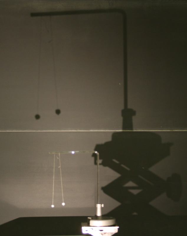 Shadow projection of the pith ball apparatus on a wall, with the actual illuminated apparatus visible in the foreground below the enlarged shadow