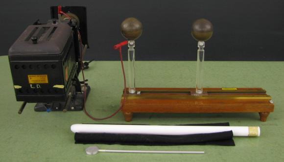 Projection electroscope connected by red alligator clip lead to one of two copper spheres on Teflon supports on a wooden base; white charging rod, black felt pad, and proof plane in foreground
