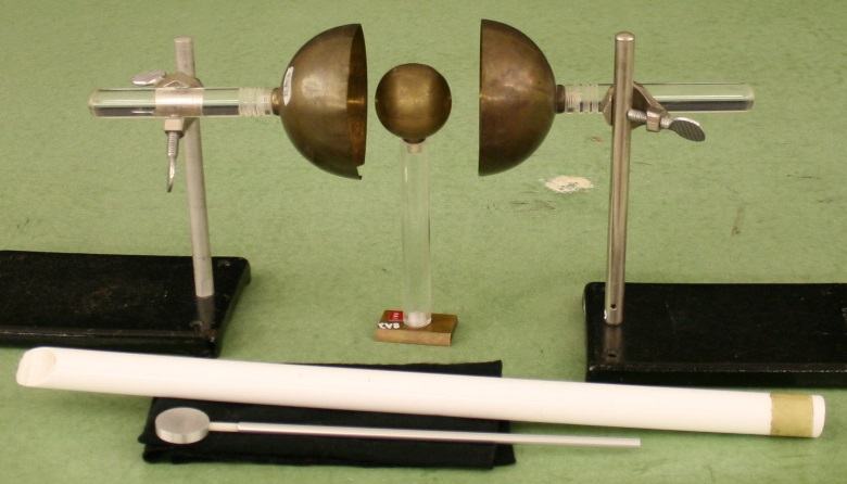 Two brass hemispheres on horizontal rods in cross clamps on black ring stands, flanking a small brass sphere on a glass insulating stand; proof plane, charging rod, and charging pad in foreground