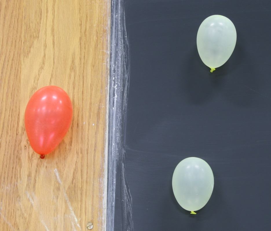 One red balloon adhering to a wooden door frame and two clear balloons stuck to a chalkboard surface, demonstrating electrostatic adhesion