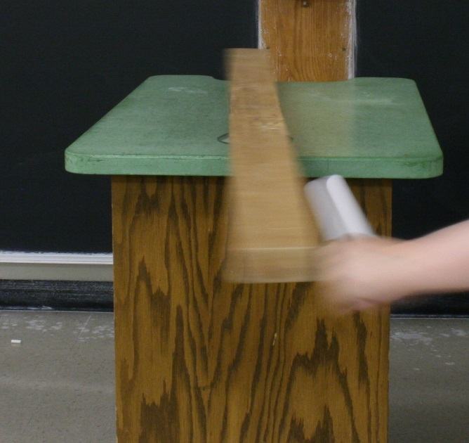 Blurred 2×4 rotating on green platform atop wooden stand, hand holding white charging rod near one end of the board