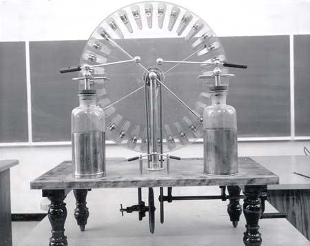 Wimshurst machine with two overlapping glass disks bearing metal sectors, two large Leyden jars, adjustable spherical discharge terminals, diagonal neutralizer bars, and hand crank on a wooden platform with turned legs