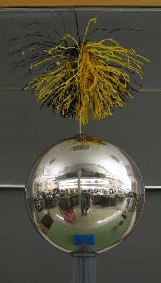 Black and gold tassel on a metal rod atop a Van de Graaff dome, with strands splayed radially outward due to electrostatic repulsion