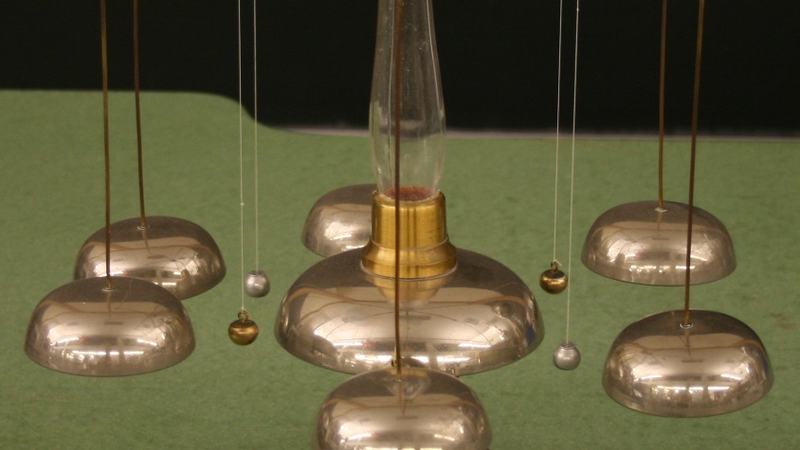 Franklin bells apparatus on a wooden base: central glass column with brass fitting and radiating brass arms, six polished dome-shaped bells in three pairs, with small metal clappers suspended on threads between the central column and each outer bell