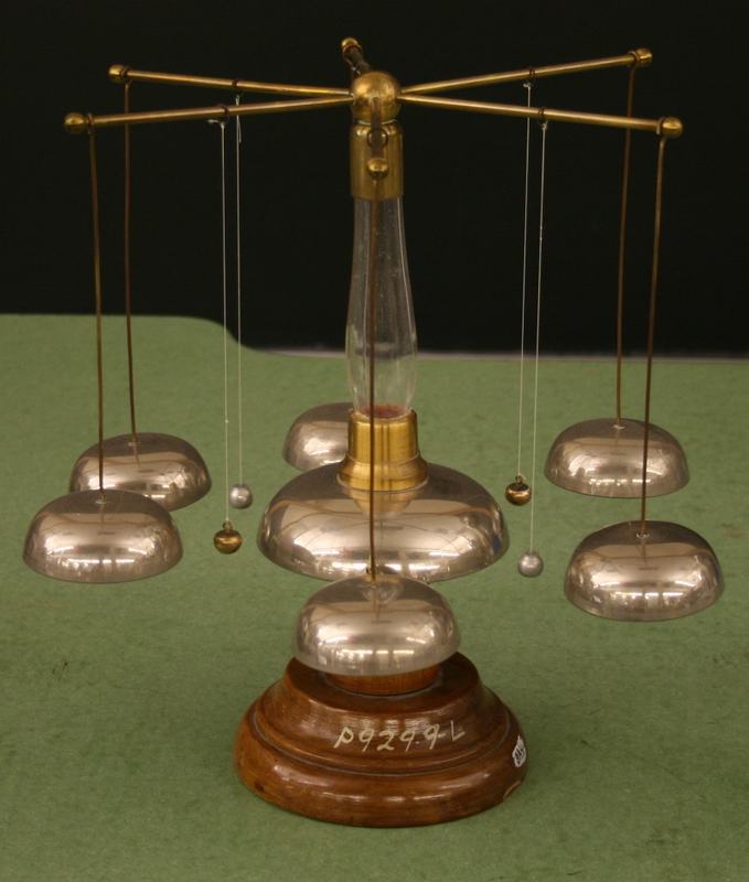 Franklin bells apparatus on a wooden base: central glass column with brass fitting and radiating brass arms, six polished dome-shaped bells in three pairs, with small metal clappers suspended on threads between the central column and each outer bell