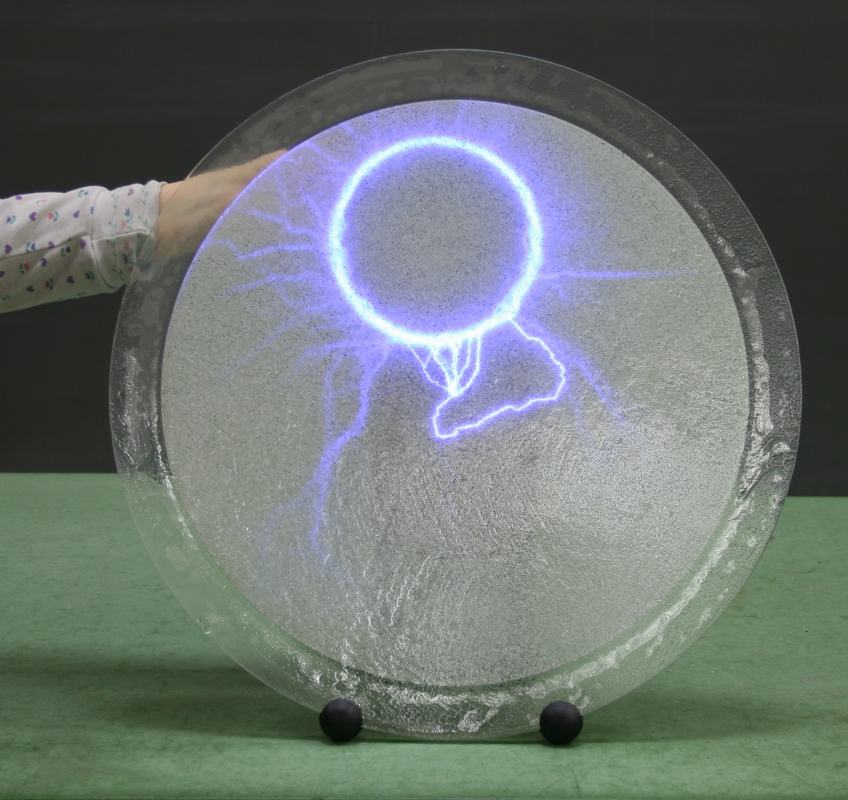 Plasma disk with metal ring in place; a hand touches the back and discharge paths form a bright circle at the ring edge, with streamers branching around but not crossing the interior