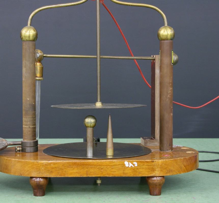 Close-up of brass lightning apparatus showing pointed cone electrode (left) and ball-topped electrode (right) on base plate beneath horizontal brass disc