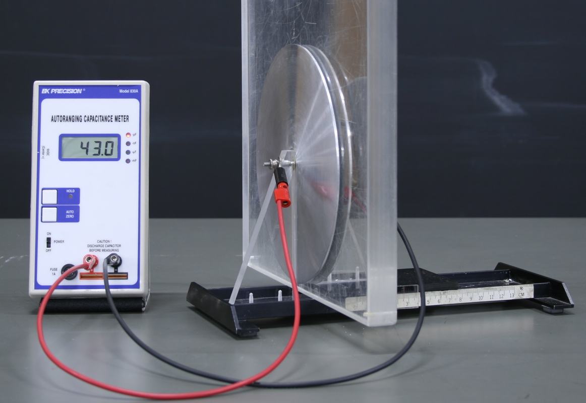 Parallel plate capacitor with circular metal plates mounted vertically on a rail base, connected via red and black leads to a BK Precision Model 830A autoranging capacitance meter reading 43.0 pF