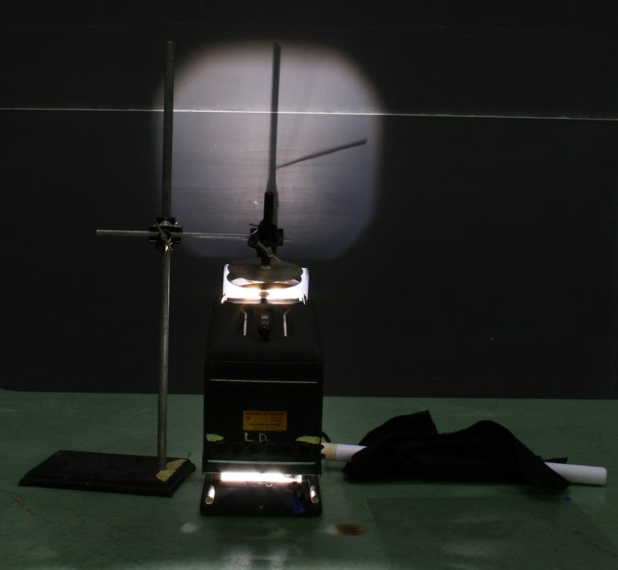 Darkened room with projection electroscope illuminated, leaf shadow projected on board showing deflection; charging rod and fur pad visible on bench