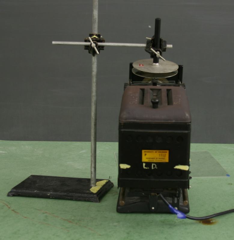 Projection electroscope with capacitor plates mounted on top via cross clamps and rod, ring stand to the left, on green bench surface