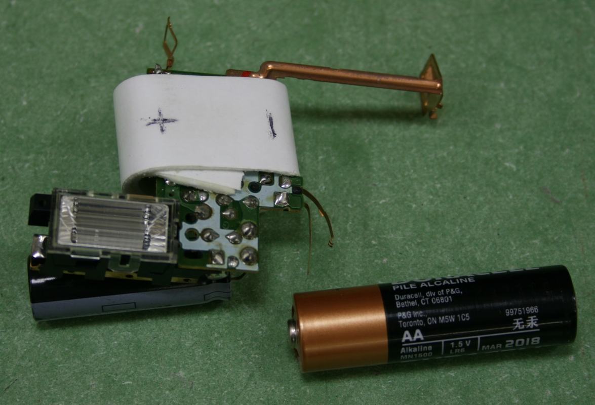 Bare circuit board from a disposable camera flash with white cylindrical 100 μF capacitor, small xenon flash tube, copper contact bracket, and Duracell AA battery alongside