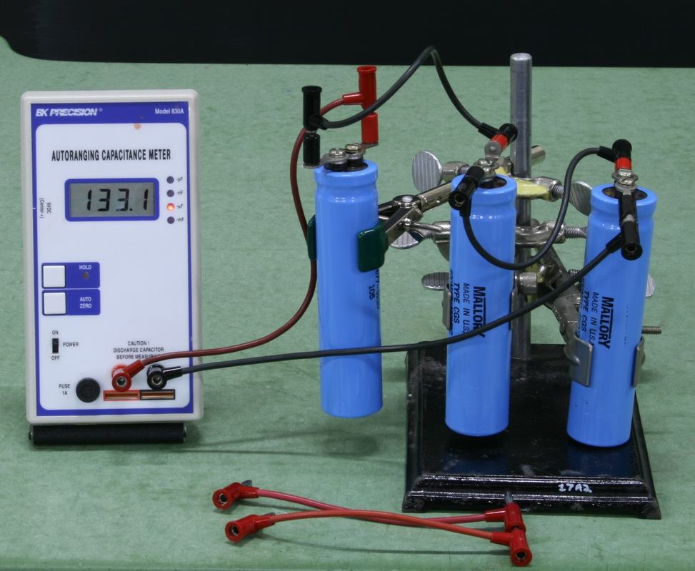 Three blue Mallory capacitors mounted vertically on a black metal stand with alligator clip connections, wired in series to a BK Precision Model 830A autoranging capacitance meter reading 133.1 µF; red jumper cables in foreground