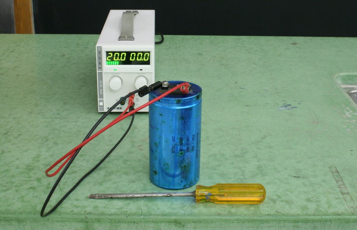 Large blue cylindrical capacitor with red bolt terminals on top, connected via red and black leads to a Xantrex DC power supply displaying 20.0V and 0.0A, with a yellow-handled screwdriver on the green table