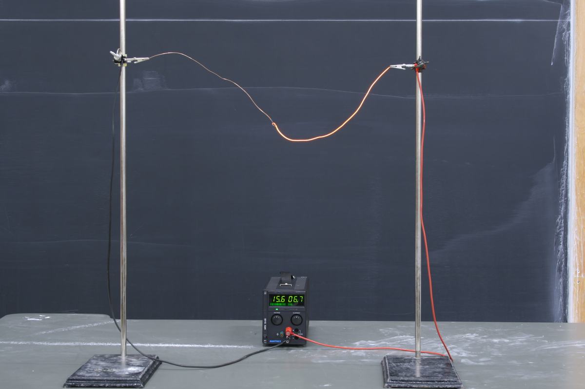 Two ring stands with right-angle clamps supporting copper and nichrome wires connected in series between them, with a DC power supply on the bench displaying 15.6V and 6.7A