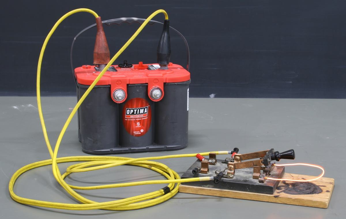 Optima car battery with red top connected by yellow cables to a wooden board holding a knife switch with thin iron wire mounted across the terminals