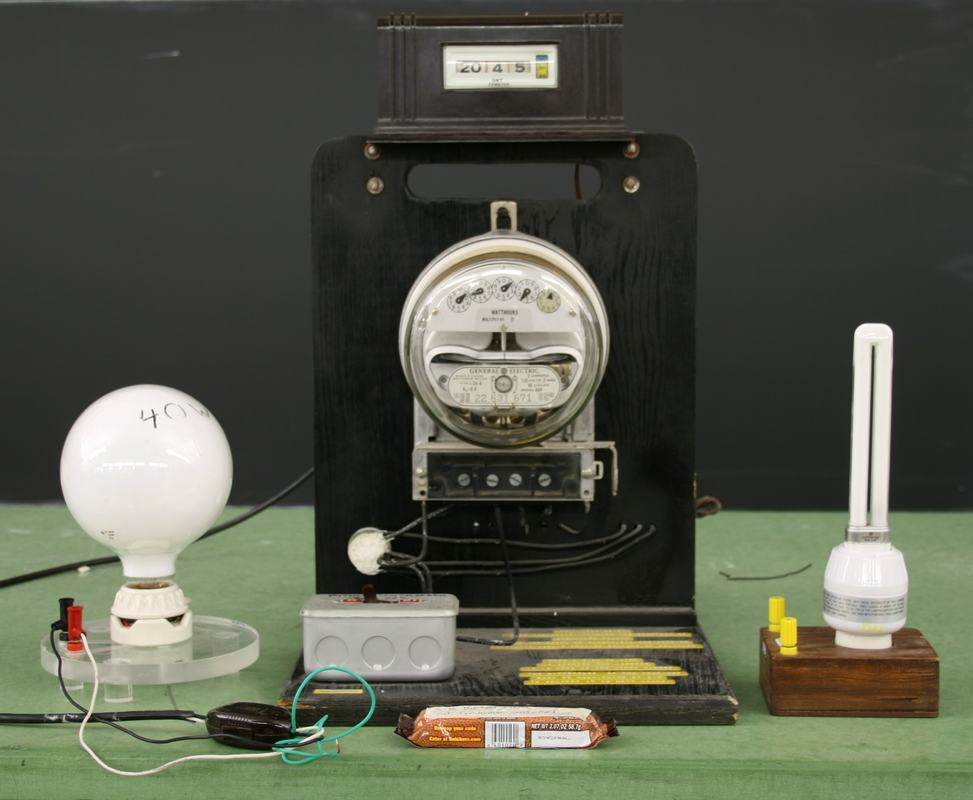 GE watthour meter mounted on a black display board with a 40W incandescent bulb on the left, control switch box in front, and a 13W compact fluorescent bulb on the right