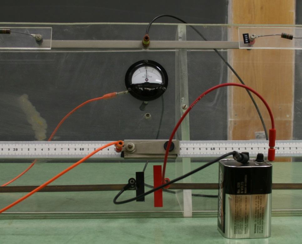 Close-up of the Wheatstone bridge showing the slide wire along a centimeter ruler, red and orange leads to the galvanometer, the sliding contact, and the Energizer battery connection