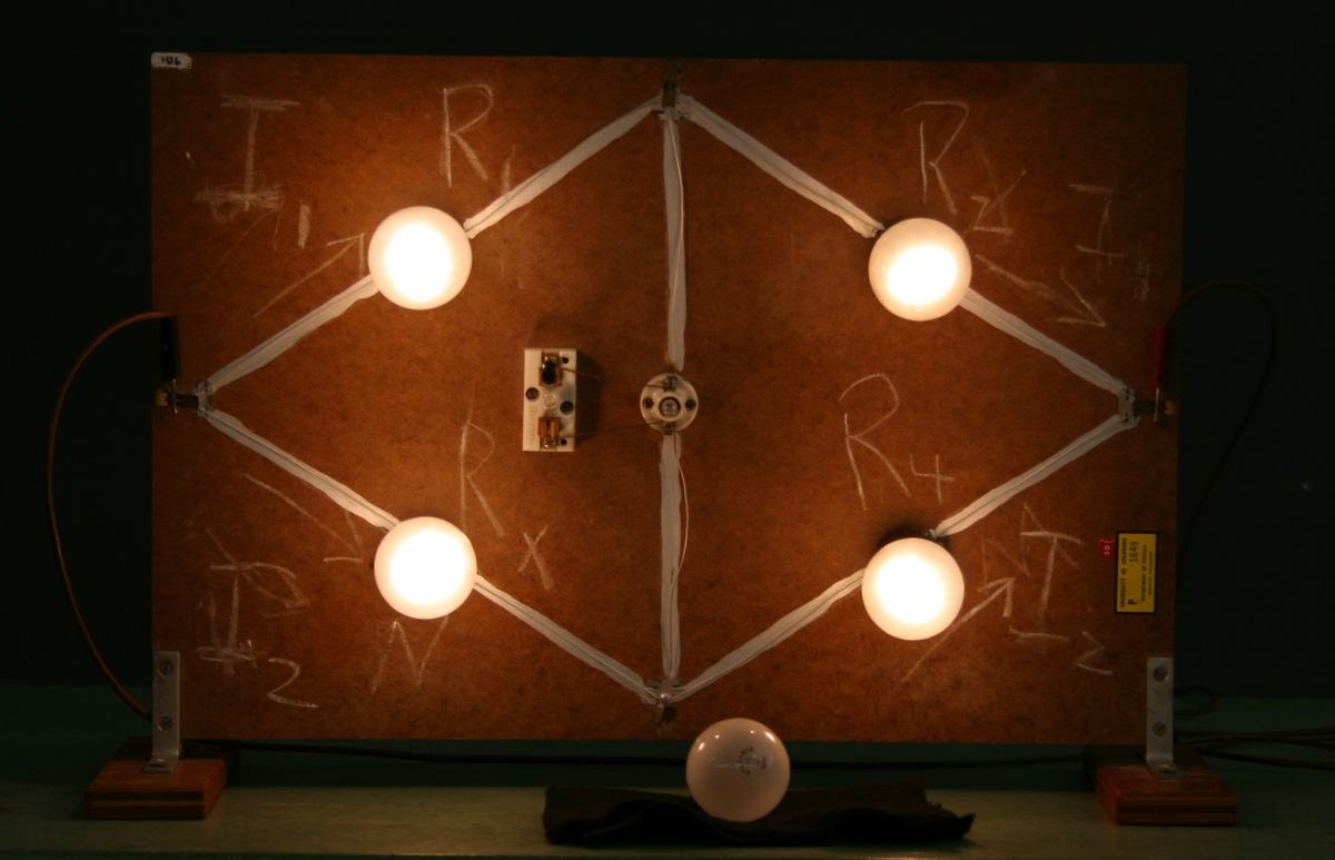 Wheatstone bridge board powered with one 60 W bulb replaced by 25 W. The four corner bulbs glow at different brightnesses and the center indicator bulb is lit, showing current through the bridge.
