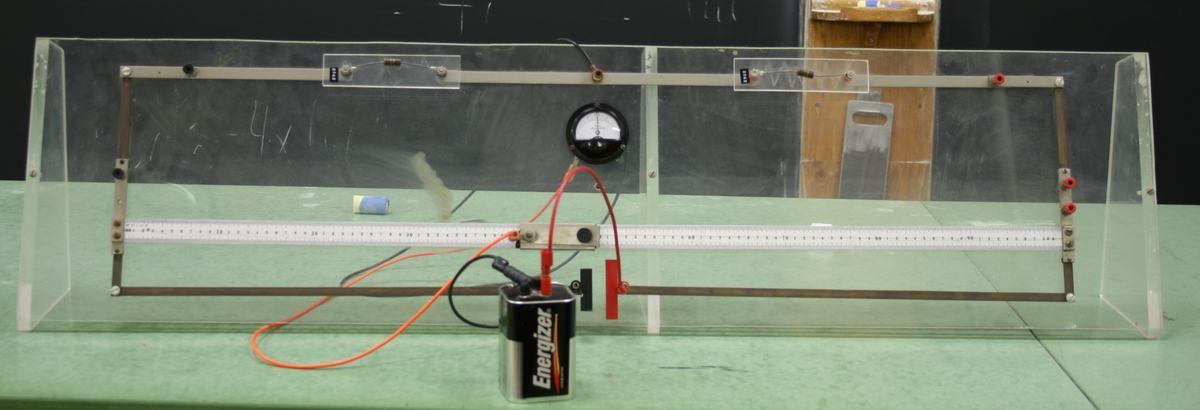 Demonstration Wheatstone bridge on a clear acrylic frame with horizontal slide wire along a centimeter ruler, center-zero galvanometer mounted above, two plug-in resistor positions at top, and an Energizer 6V battery connected below