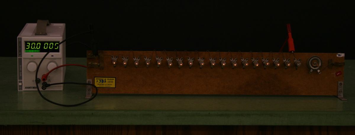 Wooden board with eighteen small incandescent bulbs in sockets wired in series, connected to a DC power supply displaying 30.0 V via red and black leads with alligator clips